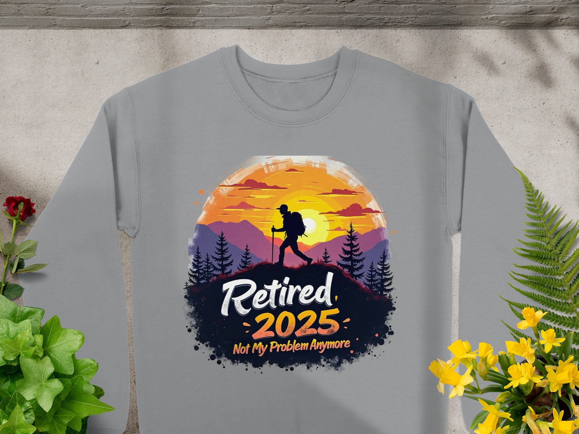 Garment Graphics offers a gray unisex sweatshirt featuring a hiker silhouette against mountains and a sunset with Retired 2025, Not My Problem Anymore. The design, surrounded by plants and flowers on concrete, caters to outdoor lovers.