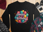 A black Happy Birthday sweatshirt by Garment Graphics features colorful balloons and stars around bold white text. Perfect for celebrations, it sits on a wooden surface with winter accessories like a hat, gloves, and plaid shirt. Available in unisex sizing.