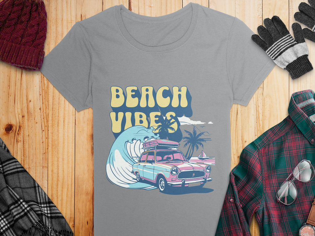 The Garment Graphics retro t-shirt showcases a surf design with a vintage car, surfboard, and palm trees. BEACH VIBES is printed on gray fabric. Styled on wood with a plaid shirt, scarf, gloves, and knitted hat for a nostalgic vibe.