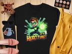 The Garment Graphics black unisex t-shirt features a cartoon character with green goggles and gloves holding a glowing green object, with the phrase Its Hero Time! Its artfully staged alongside yellow jacket, eyeshadow palette, makeup brushes, sunglasses, purse, and shoes.