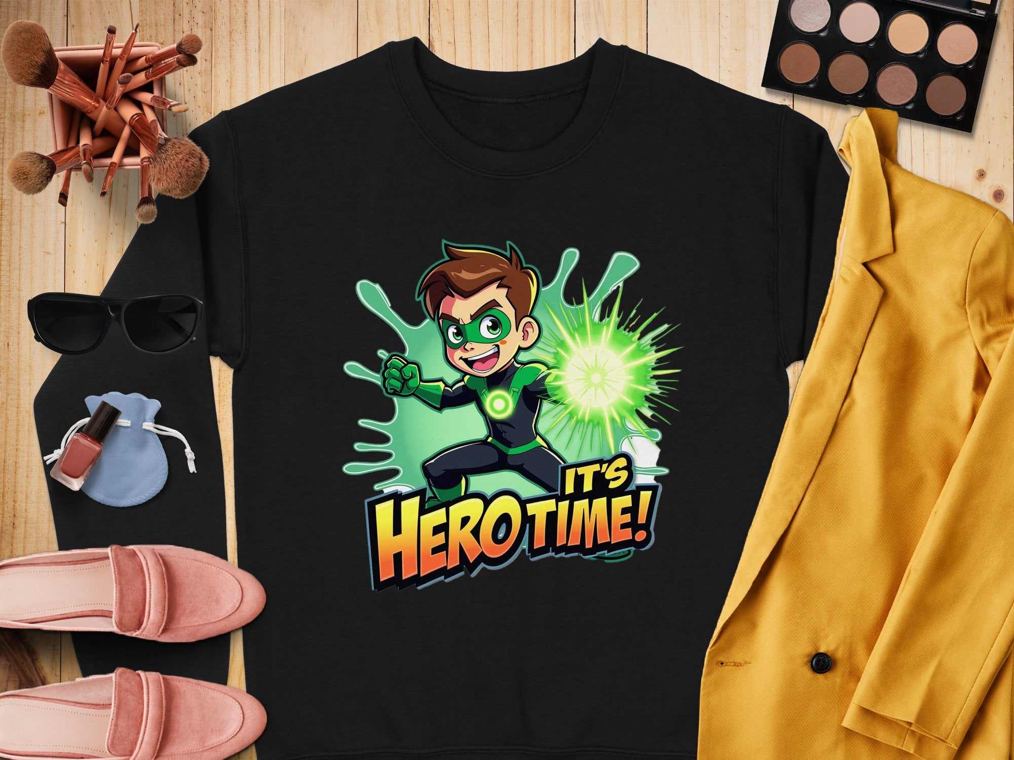 The Garment Graphics black unisex t-shirt features a cartoon character with green goggles and gloves holding a glowing green object, with the phrase Its Hero Time! Its artfully staged alongside yellow jacket, eyeshadow palette, makeup brushes, sunglasses, purse, and shoes.