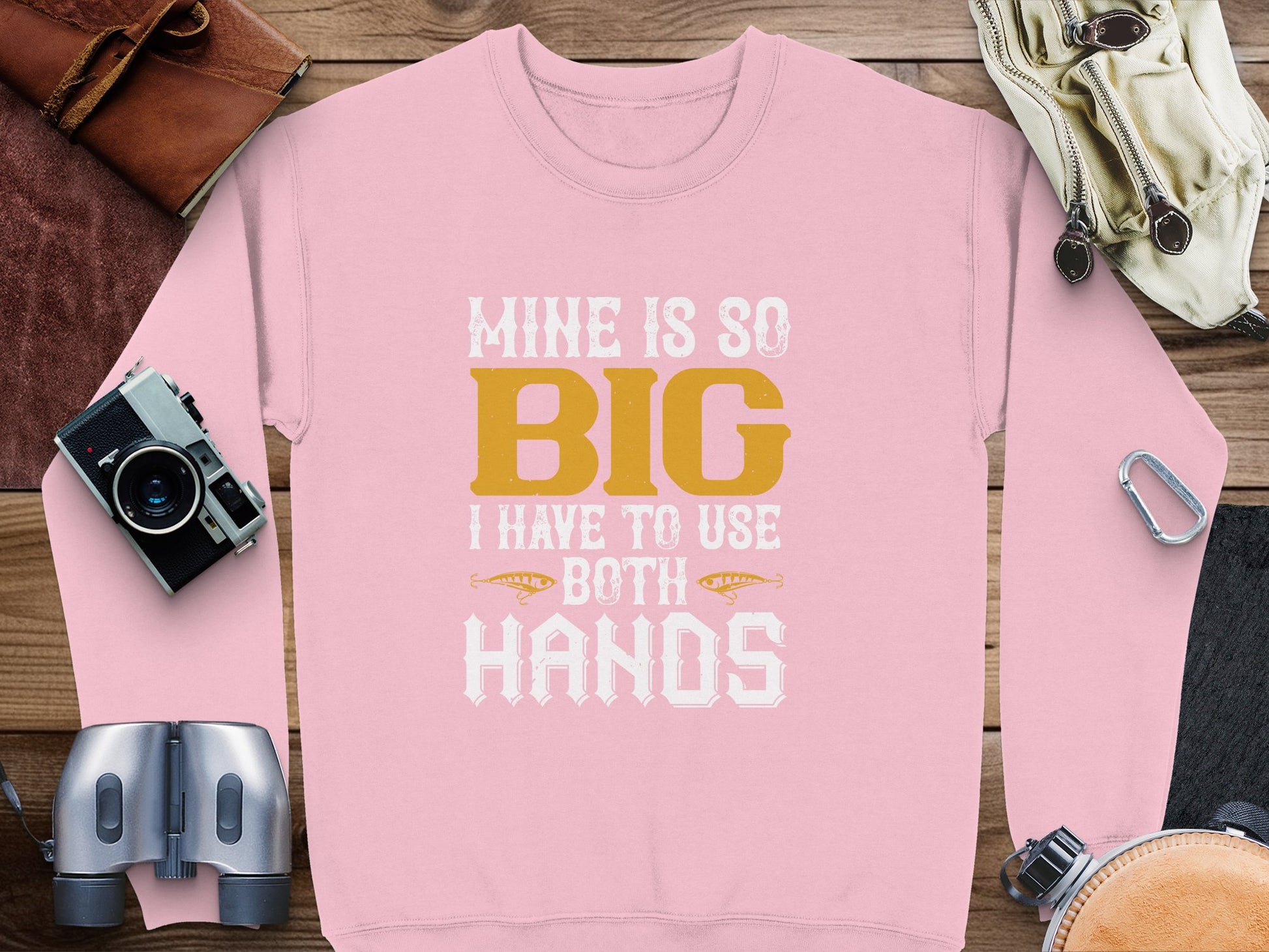 Garment Graphics offers a bold pink sweatshirt with Mine Is So Big I Have To Use Both Hands in white and yellow. It’s ideal for adventurous souls, as shown when laid on a wooden surface with binoculars, a camera, gloves, and ropes.