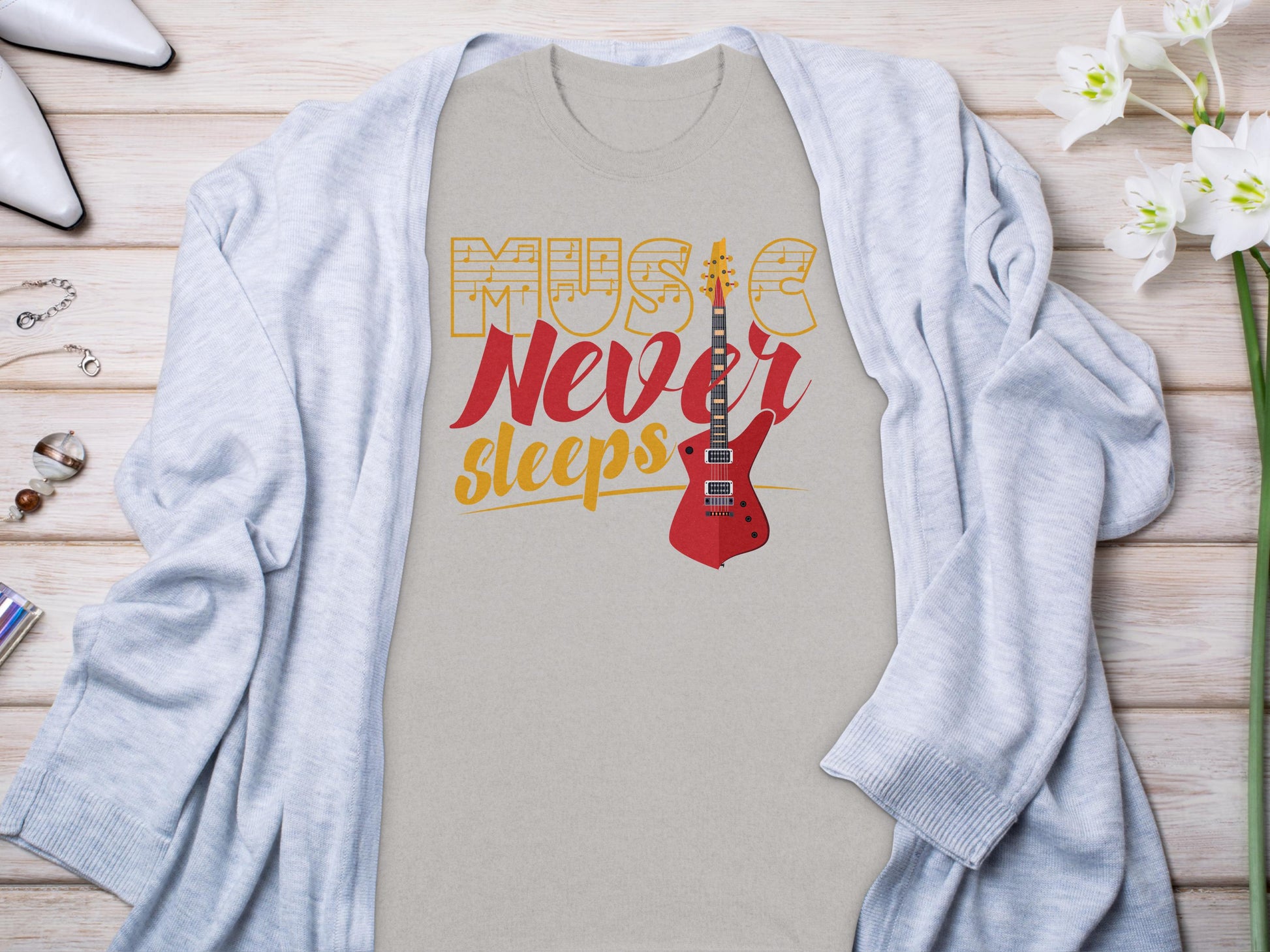 A beige Garment Graphics 100% cotton graphic t-shirt featuring Music Never Sleeps and a red electric guitar is laid flat, surrounded by a light gray cardigan, white heels, jewelry, and flowers on a wooden surface.