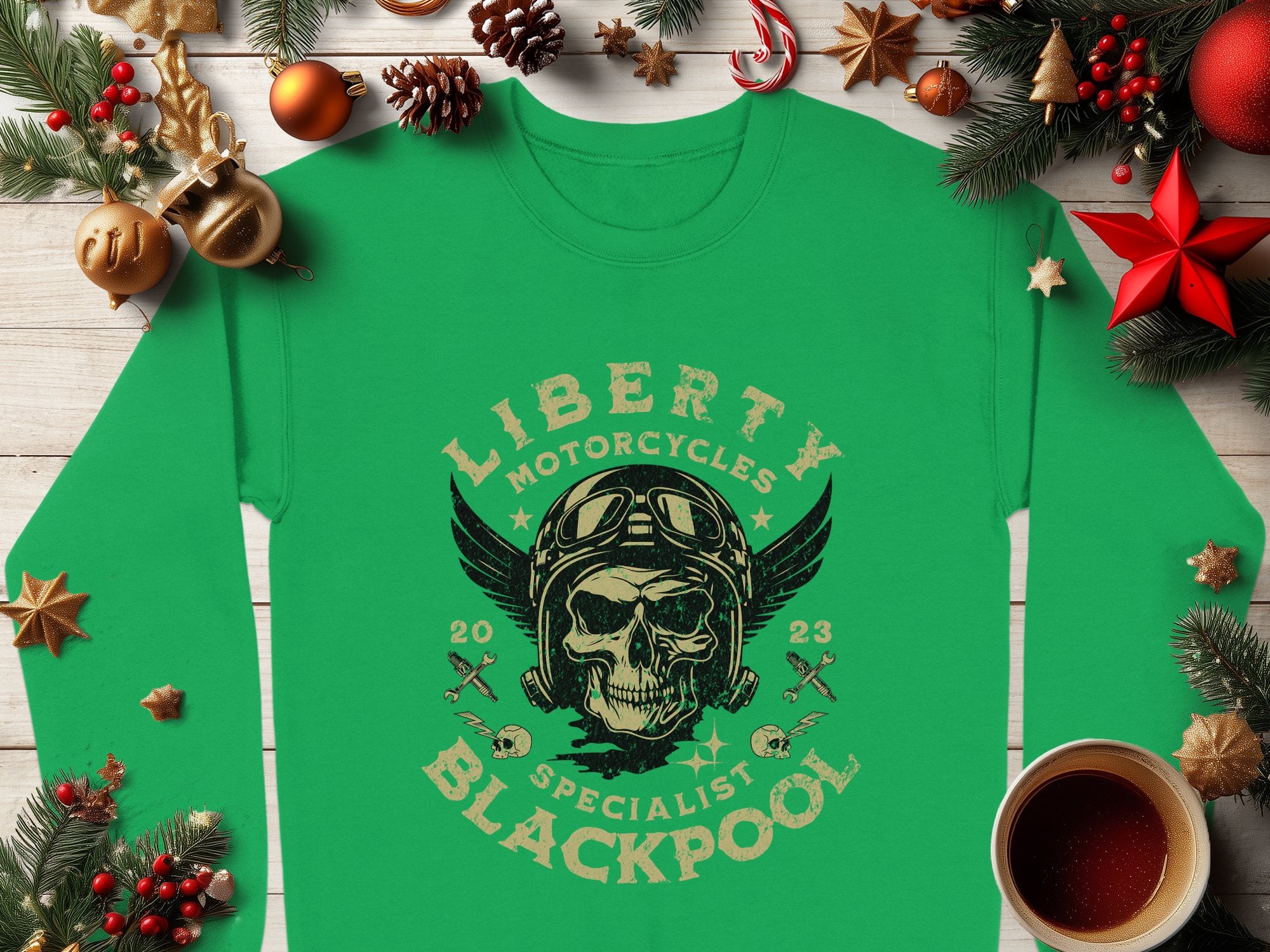 Garment Graphics offers a green shirt featuring a Liberty Motorcycles Specialist Blackpool design with a skull adorned with goggles and wings, surrounded by pine branches, ornaments, a wrapped gift, an orange, and a drink on wood.