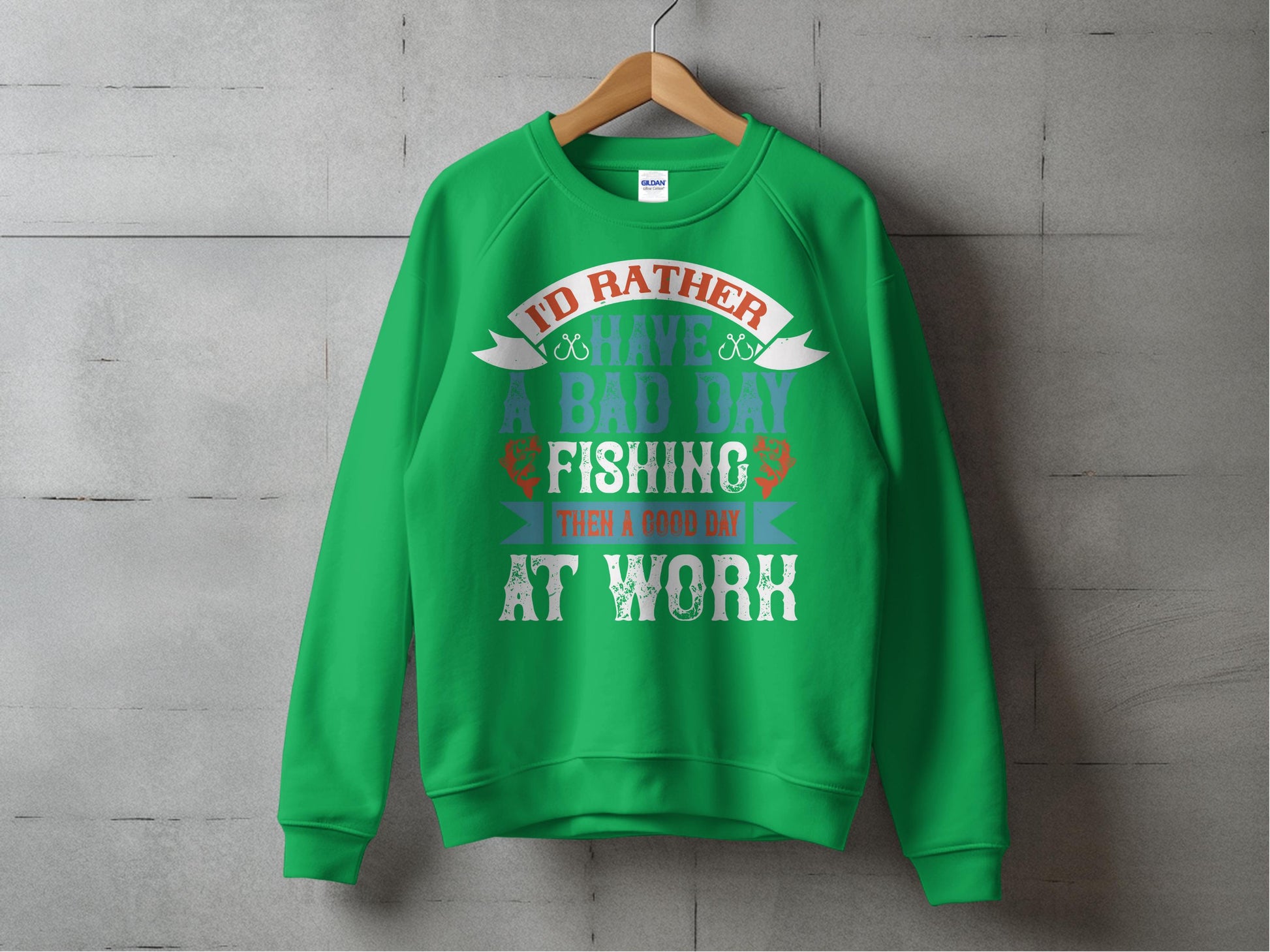 The Garment Graphics fishing sweatshirt for enthusiasts is displayed on a wooden hanger against a gray wall. This green, unisex design features playful text: Id rather have a bad day fishing than a good day at work, embellished with colorful letters and fishing-themed graphics.