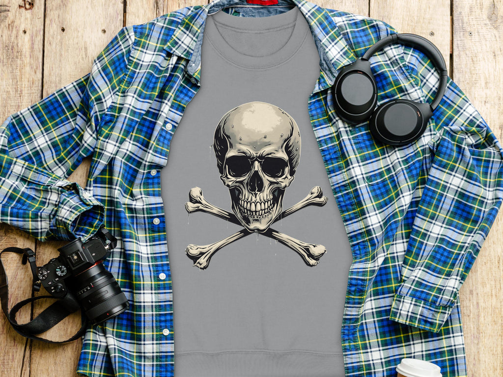 A gray Garment Graphics shirt with a skull and crossbones design, ideal for an edgy look, is displayed on a wooden surface. Nearby are a blue-green plaid shirt, black camera, headphones, and paper cup. The unisex sizing ensures its adaptable for any wardrobe.