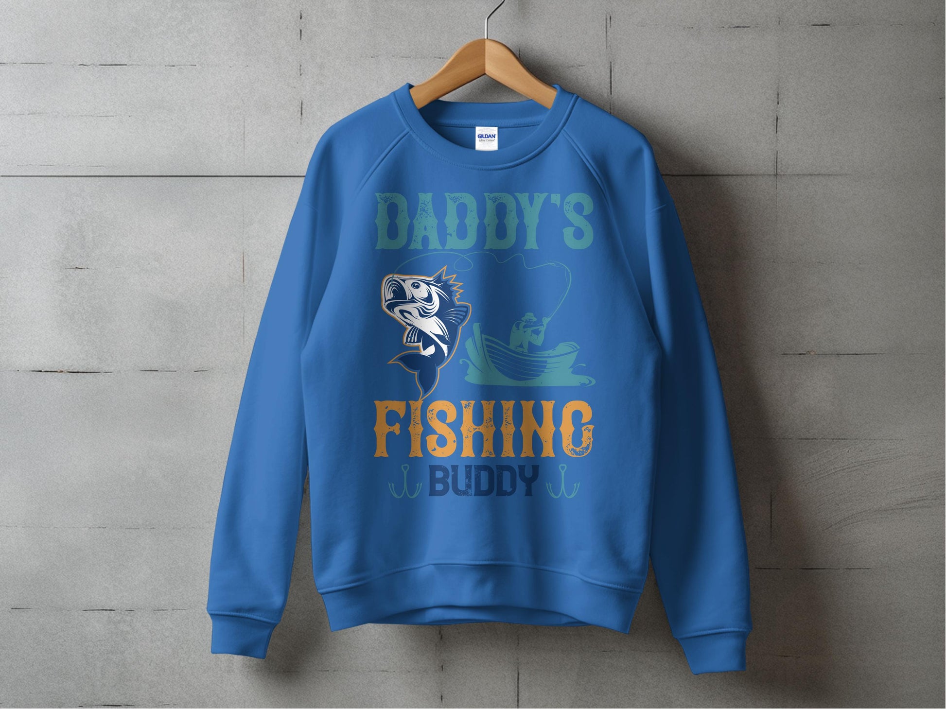 A blue fishing sweatshirt by Garment Graphics hangs on a wooden hanger against a concrete wall. It features Daddys Fishing Buddy text with a fish and person fishing from a boat, ideal for fishing enthusiasts who love spending family time on the water.