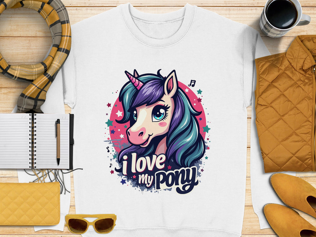 A Garment Graphics white shirt features a colorful unicorn design with music notes, stars, and the phrase I love my pony, laid out on wood with a notebook, pen, scarf, cup, and yellow accents.