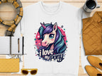 A Garment Graphics white shirt features a colorful unicorn design with music notes, stars, and the phrase I love my pony, laid out on wood with a notebook, pen, scarf, cup, and yellow accents.