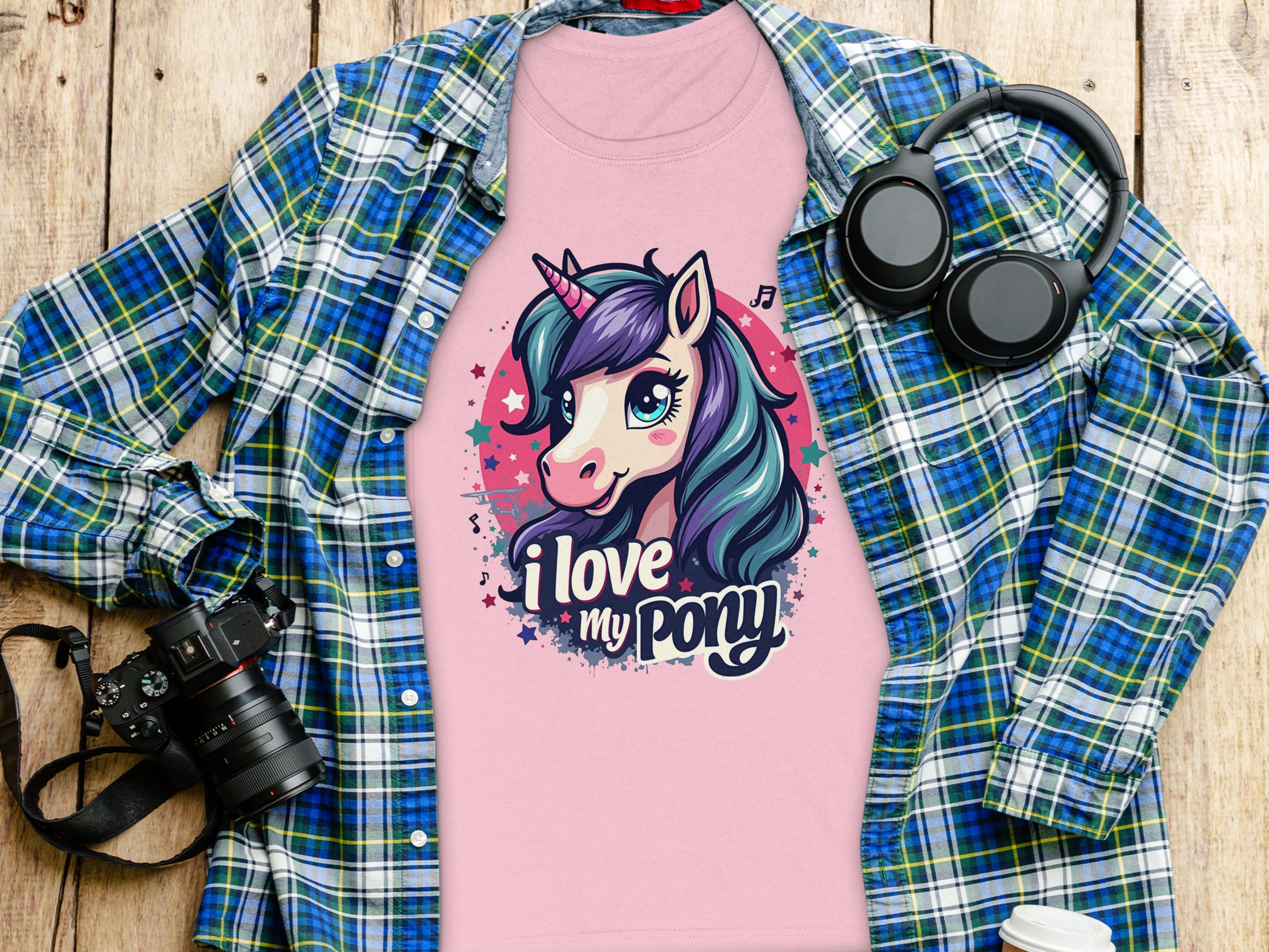 A whimsical pink T-shirt from Garment Graphics, ideal for unicorn lovers, features a unicorn graphic with I love my pony text. Its creatively displayed on wood alongside a blue and green flannel shirt, camera, headphones, and a coffee cup.