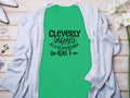 A Garment Graphics green Cleverly Disguised as a Responsible Adult T-Shirt with black text rests on a wooden surface, draped in a light gray cardigan—ideal for casual outings. Style it with white shoes and silver jewelry from the glass jar for a classic look.