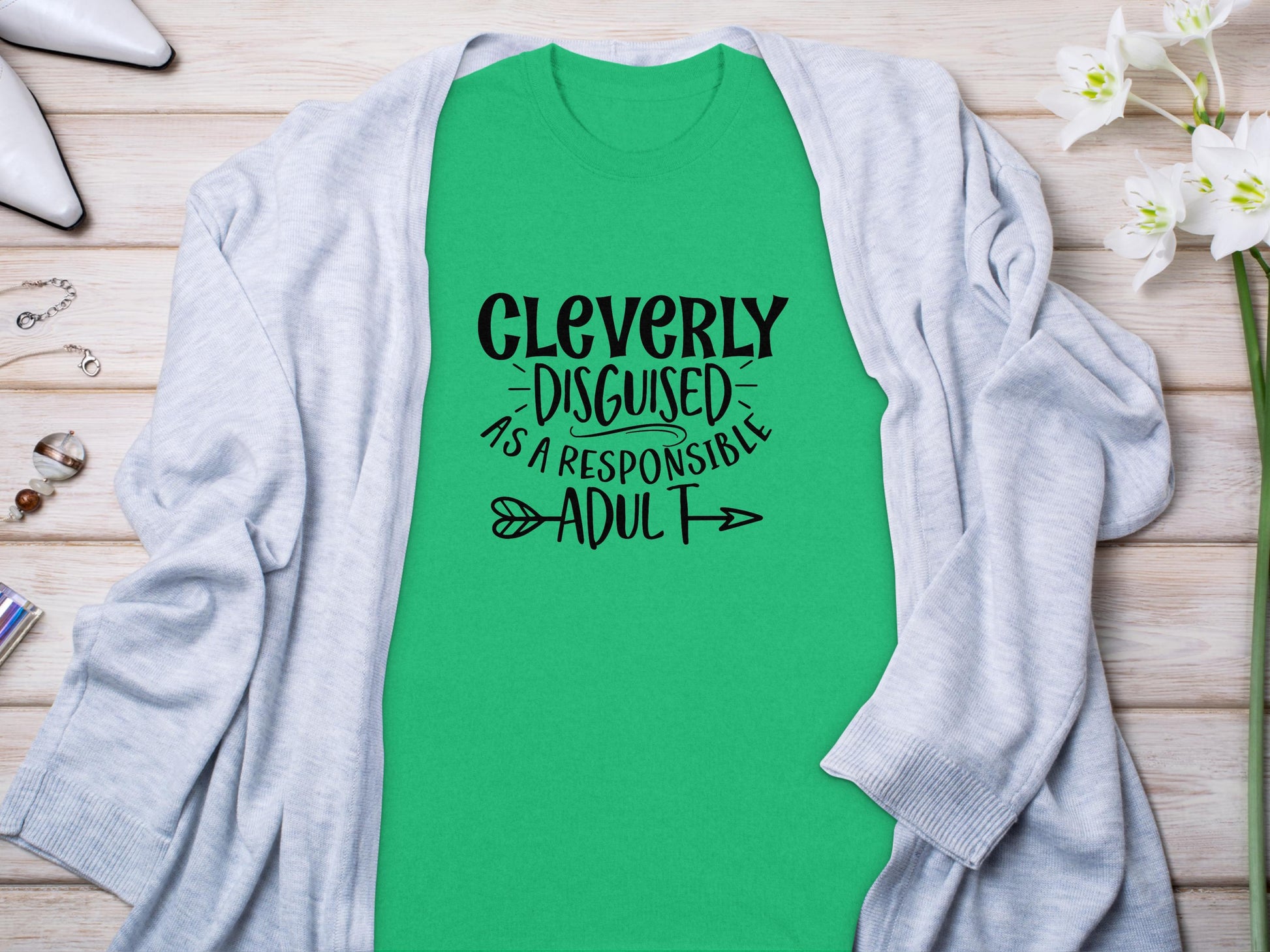 A Garment Graphics green Cleverly Disguised as a Responsible Adult T-Shirt with black text rests on a wooden surface, draped in a light gray cardigan—ideal for casual outings. Style it with white shoes and silver jewelry from the glass jar for a classic look.