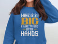 The woman wears a vibrant blue Garment Graphics sweatshirt with MINE IS SO BIG I HAVE TO USE BOTH HANDS in bold white and yellow font. Adjusting her hair, the statement piece enhances her casual look.