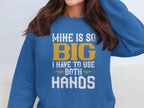 The woman wears a vibrant blue Garment Graphics sweatshirt with MINE IS SO BIG I HAVE TO USE BOTH HANDS in bold white and yellow font. Adjusting her hair, the statement piece enhances her casual look.