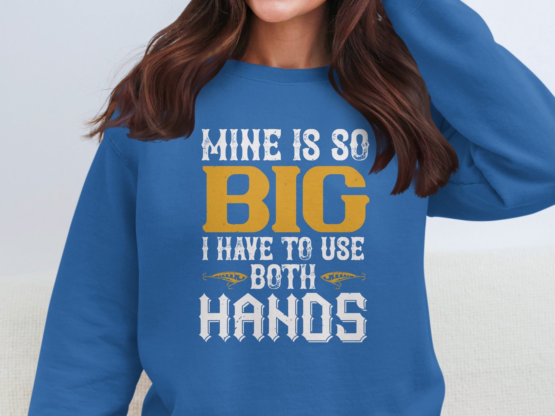 The woman wears a vibrant blue Garment Graphics sweatshirt with MINE IS SO BIG I HAVE TO USE BOTH HANDS in bold white and yellow font. Adjusting her hair, the statement piece enhances her casual look.