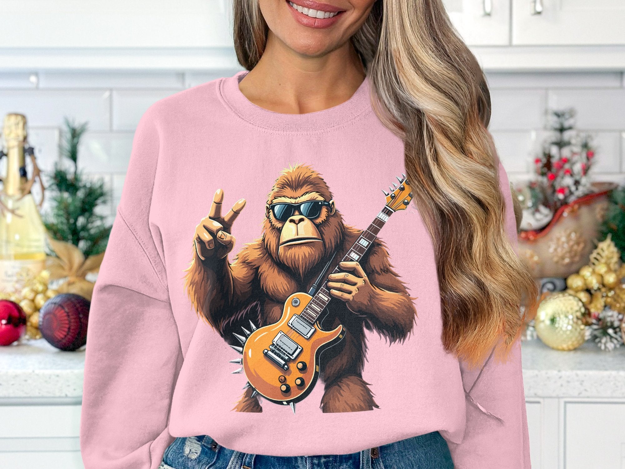 A woman with long blonde hair dons a Garment Graphics pink sweatshirt featuring a rocking gorilla illustration, playing an electric guitar and making a peace sign, set against holiday decorations like ornaments and a small tree – ideal for music lovers.