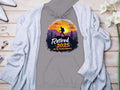 Garment Graphics gray sweatshirt features “Retired 2025: Not My Problem Anymore” in front with a sunset mountain design and hiker silhouette against a vibrant sky, showcased on wooden surface alongside a light cardigan, flowers, and accessories.