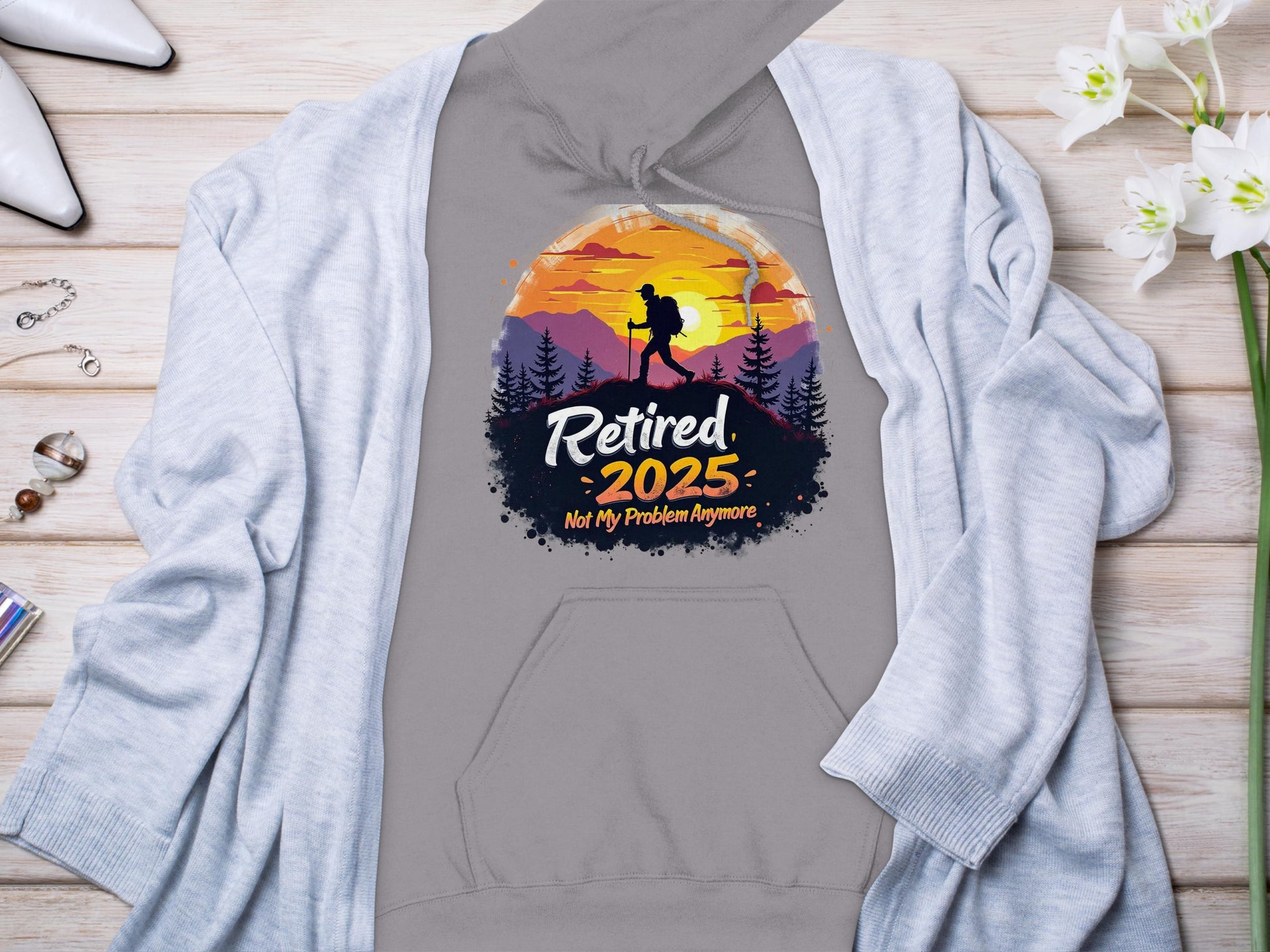 Garment Graphics gray sweatshirt features “Retired 2025: Not My Problem Anymore” in front with a sunset mountain design and hiker silhouette against a vibrant sky, showcased on wooden surface alongside a light cardigan, flowers, and accessories.