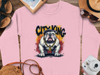 Garment Graphics presents a pink bulldog sweatshirt featuring a stylish bulldog with sunglasses and a gold chain, labeled CITY KING. Its creatively showcased on wood and accompanied by summer essentials like sunglasses and a hat.