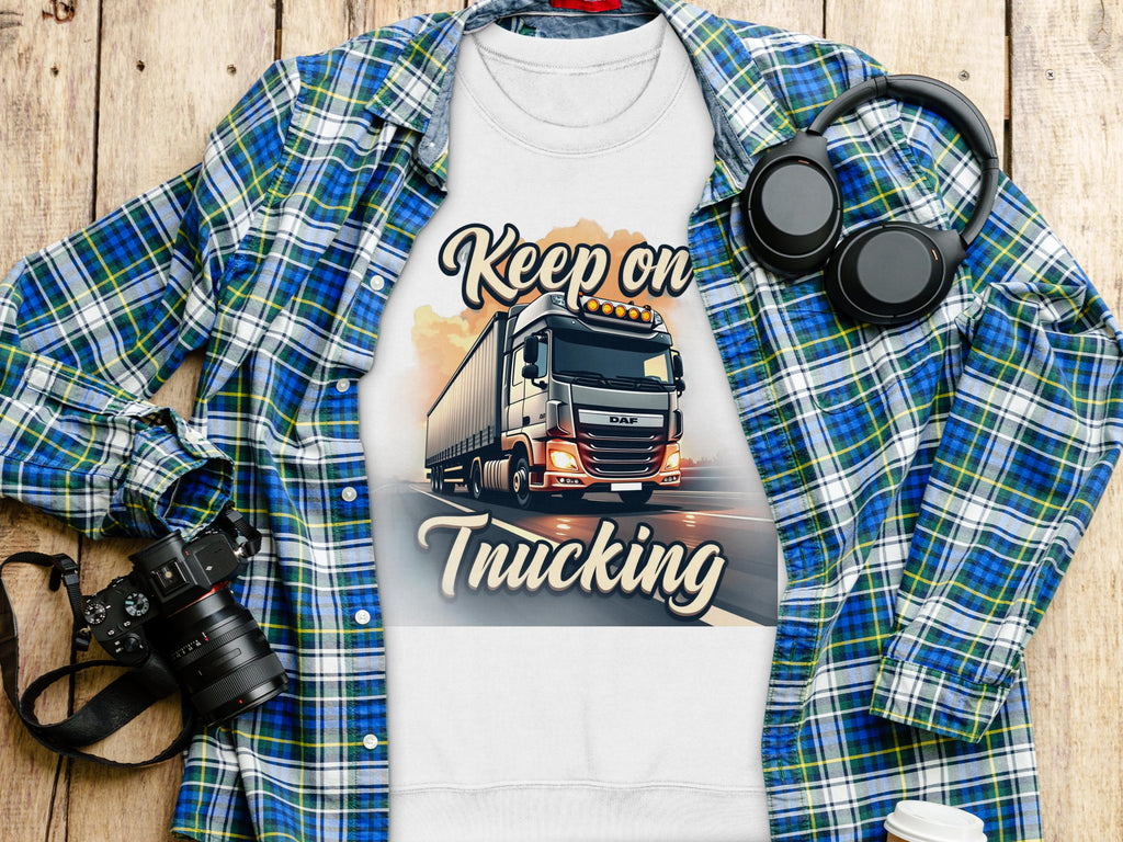 A flat lay features a Garment Graphics white T-shirt with a truck graphic and Keep on Trucking text. It embodies open-road spirit, resting on a blue and green plaid shirt, accompanied by a camera, headphones, and disposable coffee cup on the wooden surface.