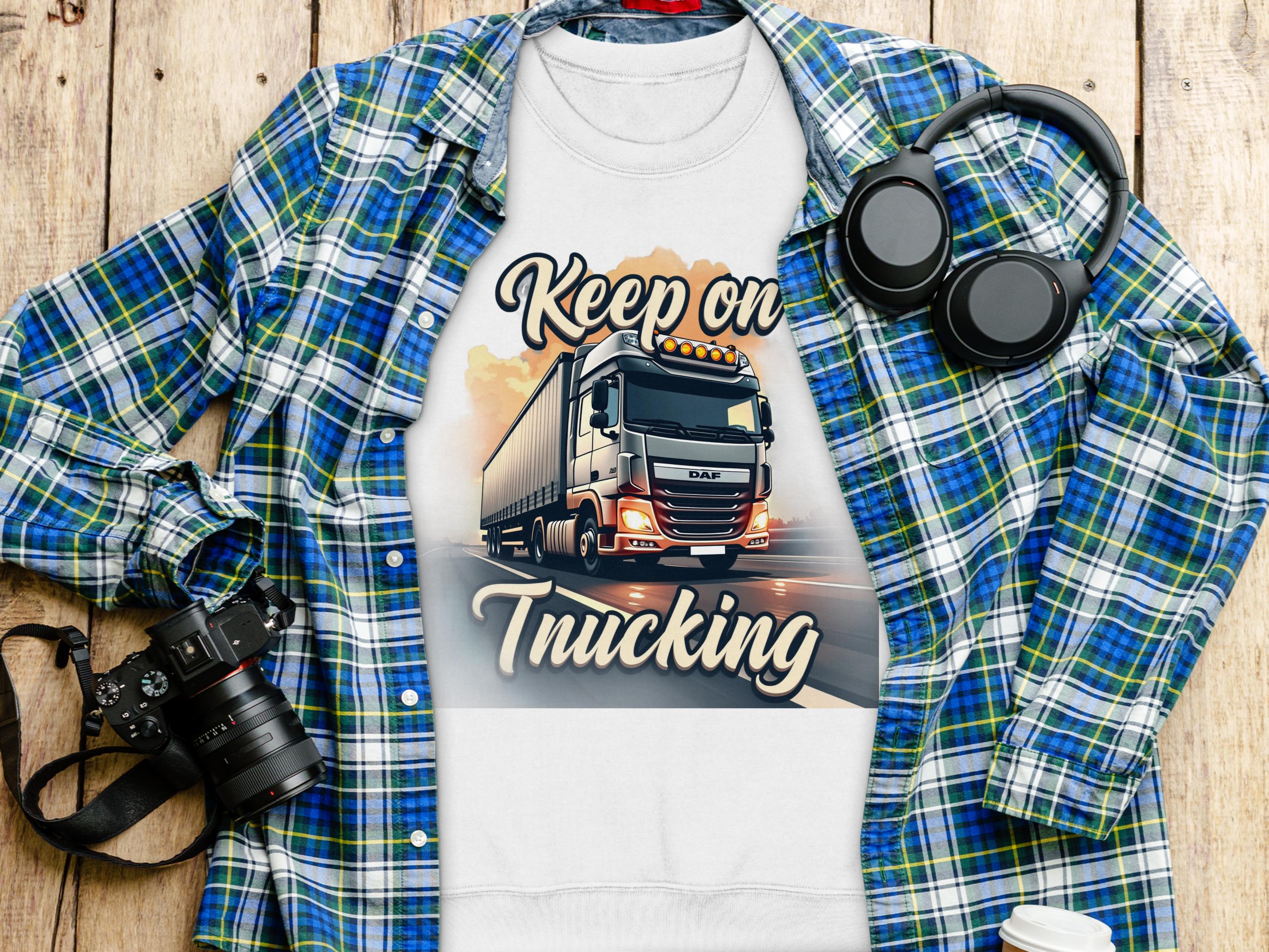 A flat lay features a Garment Graphics white T-shirt with a truck graphic and Keep on Trucking text. It embodies open-road spirit, resting on a blue and green plaid shirt, accompanied by a camera, headphones, and disposable coffee cup on the wooden surface.