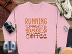 The Garment Graphics pink graphic sweatshirt, with Running on Grace & Coffee in orange and brown letters, is unisex for comfort. Its artfully displayed on a wooden surface accompanied by a camera, binoculars, leather gloves, and a coiled rope for an adventurous vibe.