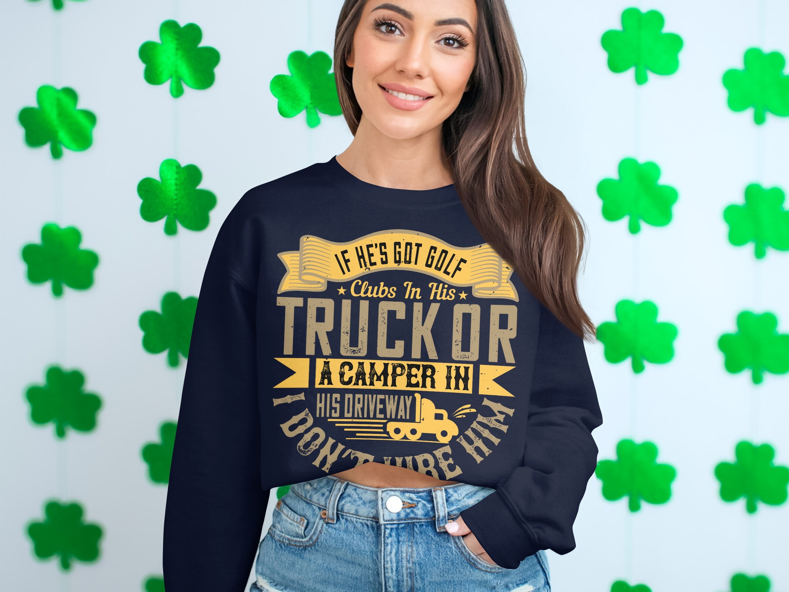 Wearing Garment Graphics humorous navy blue sweatshirt featuring the phrase, If hes got golf clubs in his truck or a camper in his driveway, I dont trust him, a person stands against a wall of green shamrocks, smiling broadly and enhancing the casual wear vibe.