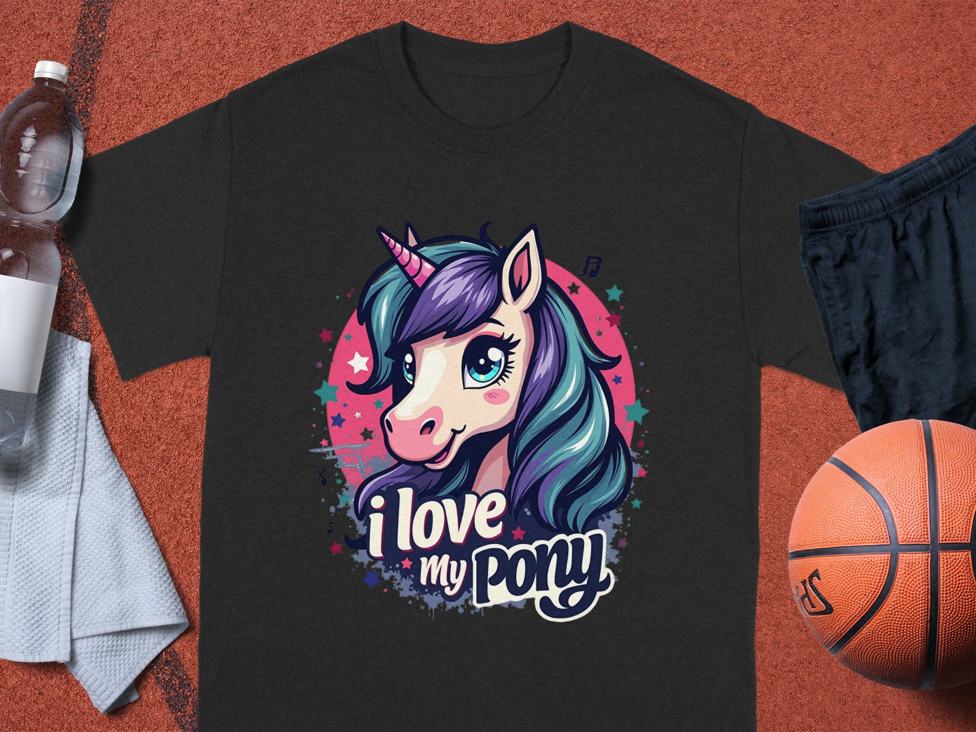 A black Garment Graphics T-shirt features a cartoon unicorn with a purple-blue mane, pink horn, and cheerful expression. The i love my pony text makes it ideal for unicorn fans. Its shown on an athletic surface with a towel, basketball, and water bottle.