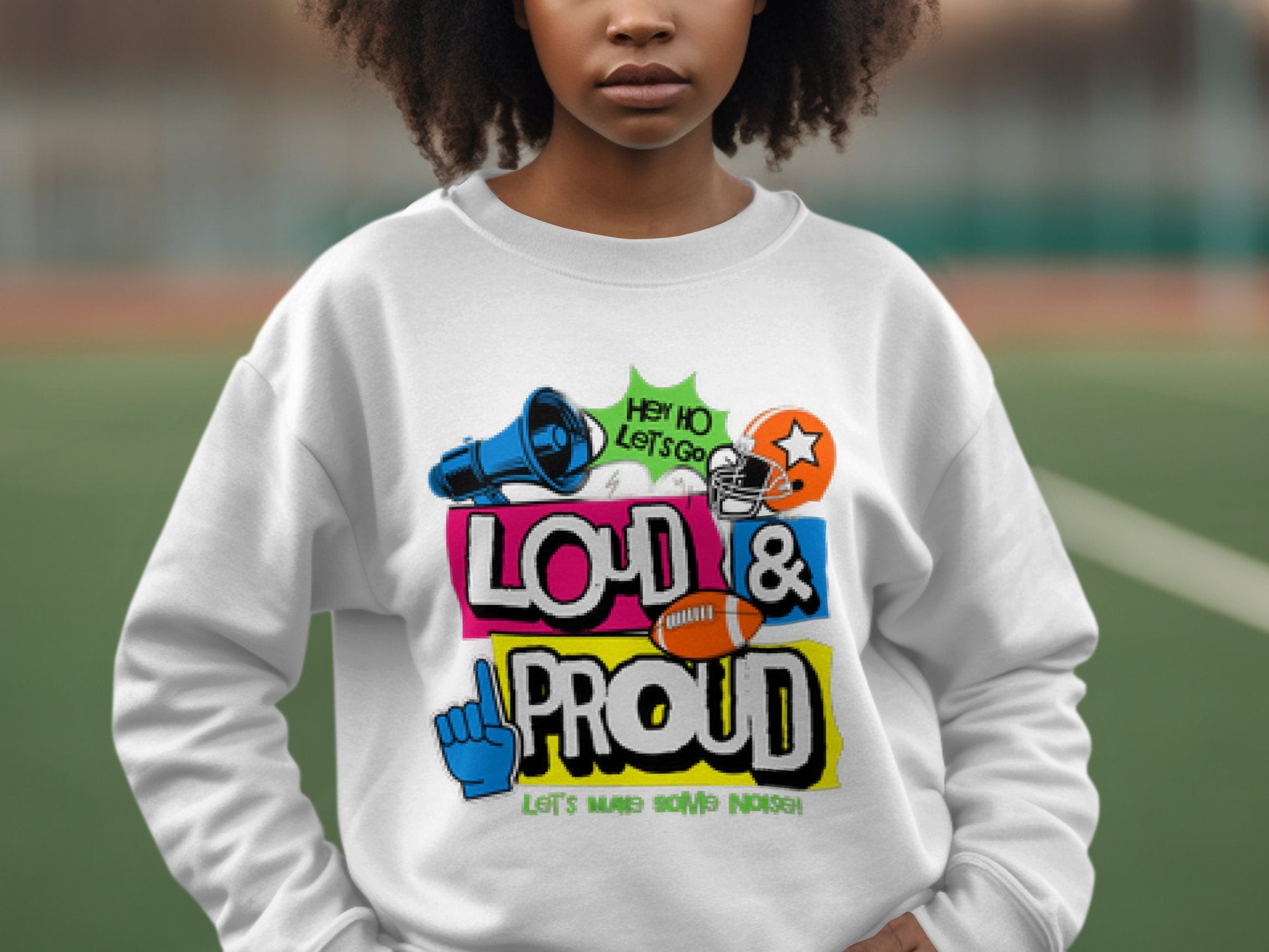 A person outdoors wears a Garment Graphics Loud and Proud white sweatshirt with unisex sizing, featuring vibrant graphics of a megaphone, football helmet, and football with the text LOUD & PROUD and Lets Make Some Noise, against a blurred background.