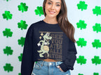 A person with long hair wears a Garment Graphics alligator sweatshirt featuring a cartoon dinosaur playing basketball and the text See You Later Alligator. Theyre standing against a light wall decorated with green shamrocks.