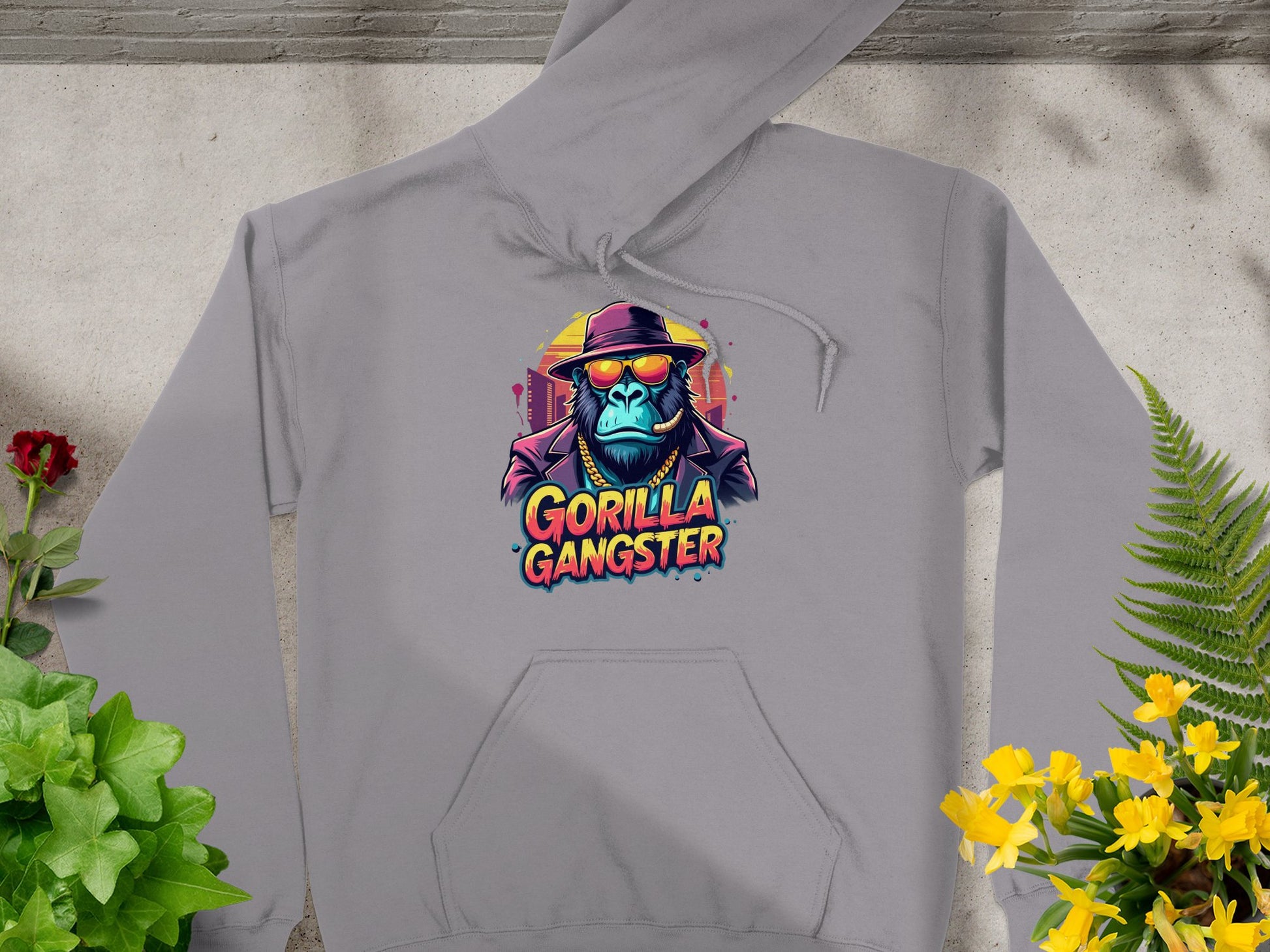 The Garment Graphics gray hoodie showcases gorillas with hats and sunglasses, reading Gorilla Gangster. Surrounded by plants and flowers on a stone surface, its a standout piece in streetwear gangster hoodies.