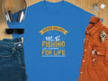 A blue Garment Graphics T-shirt with Father and Son Fishing Partners for Life in yellow and white celebrates family bonding, featuring two people fishing. Its styled with sunglasses, headphones, denim jeans, a watch, and a brown shirt on a wooden surface.