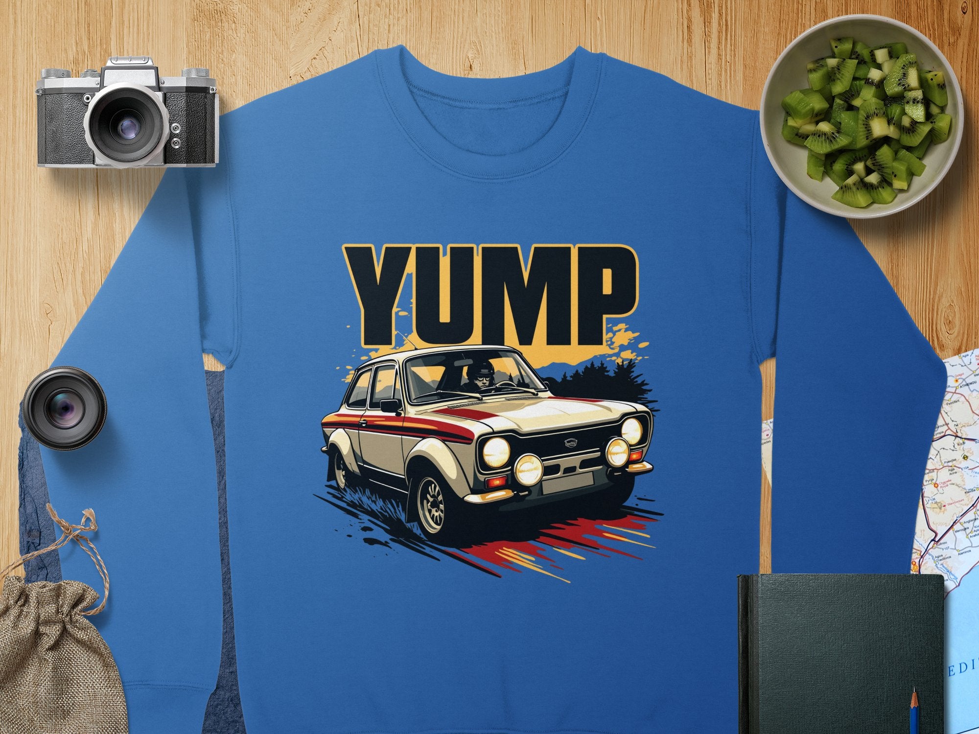 This blue sweatshirt from Garment Graphics features a striking vintage rally car design. Surrounded by essentials like a camera, lens, small bag, and green diced fruits on a wooden surface, it invites adventure and style into your wardrobe.