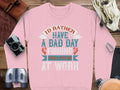 A pink Unisex Fishing Sweatshirt from Garment Graphics rests on a wooden surface, flanked by a camera, leather pouch, binoculars, coiled rope, and beige strap. It says: Id rather have a bad day fishing than a good day at work, exemplifying true Fishing Passion Apparel.