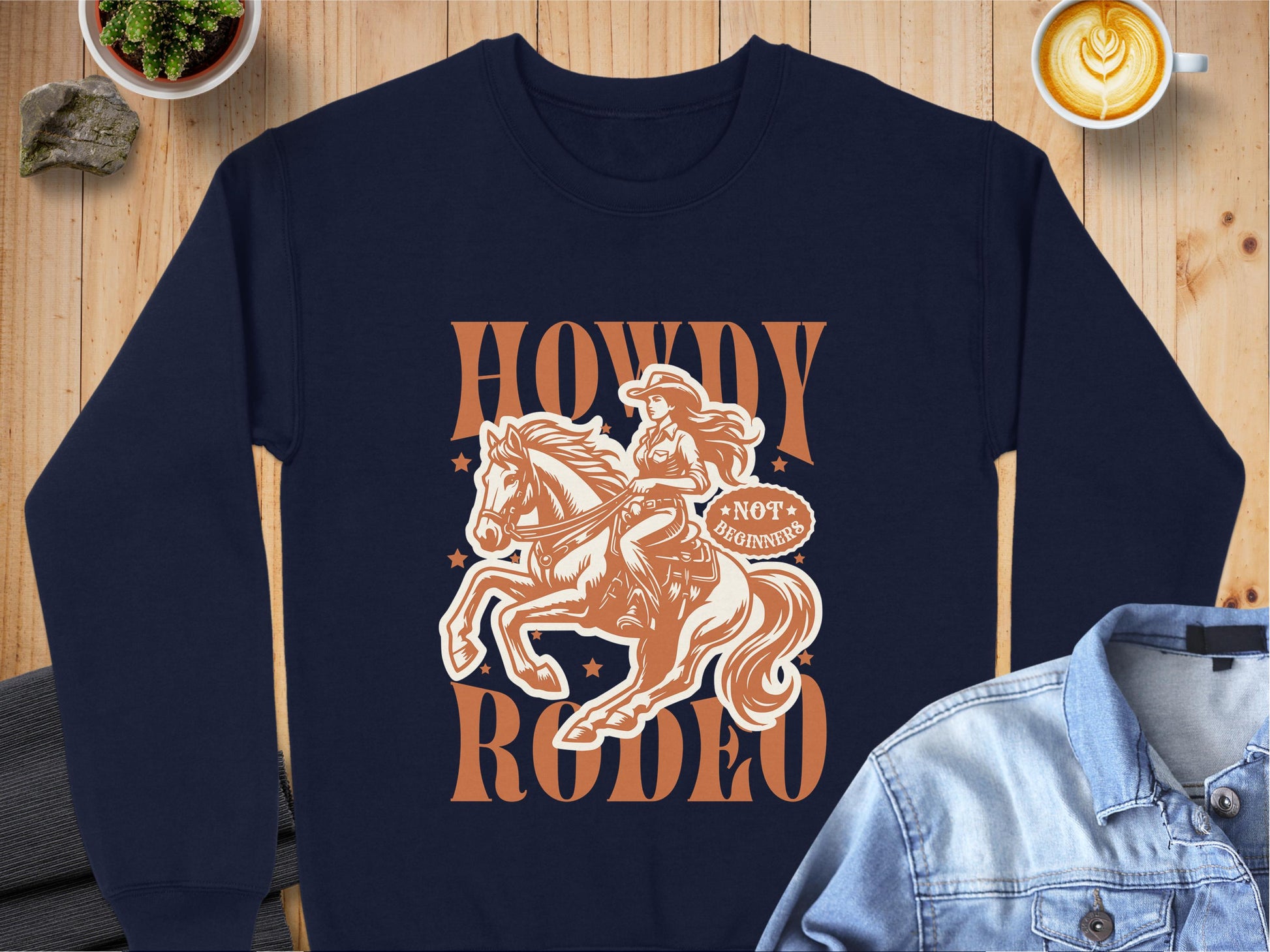 The dark blue Howdy Rodeo Sweatshirt by Garment Graphics showcases an illustrated rider on horseback amidst stars, with Howdy Rodeo and Not a Beginner phrases. Displayed on wood with a denim jacket, rocks, and a latte nearby, it embodies true cowboy spirit for rodeo enthusiasts.