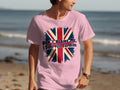 A person strolls along a beach in a pink classic fit t-shirt from Garment Graphics, showcasing a distressed Union Jack design with the bold statement, Dont blame me, I didnt vote for him, set against a backdrop of sand and ocean.