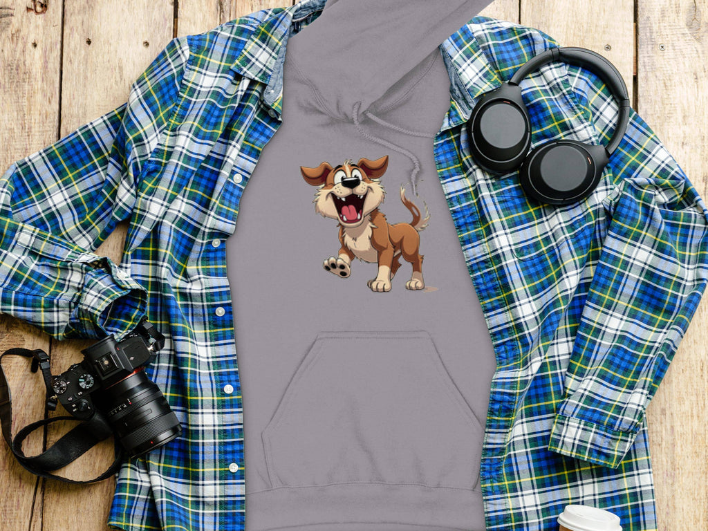 A grey cartoon puppy hoodie from Garment Graphics sits on a rustic wooden surface, accompanied by a blue and green plaid shirt, black camera, wireless headphones, and white coffee cup with lid. This stylish piece is ideal for dog lovers seeking comfort.