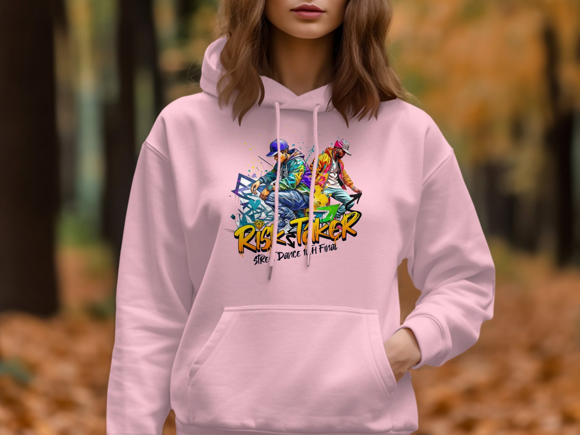 Amidst a backdrop of autumn leaves, a person wears Garment Graphics light pink hoodie. The bold design features Risk Taker Street Dance and dynamic figures, making it a statement piece for fashion enthusiasts.