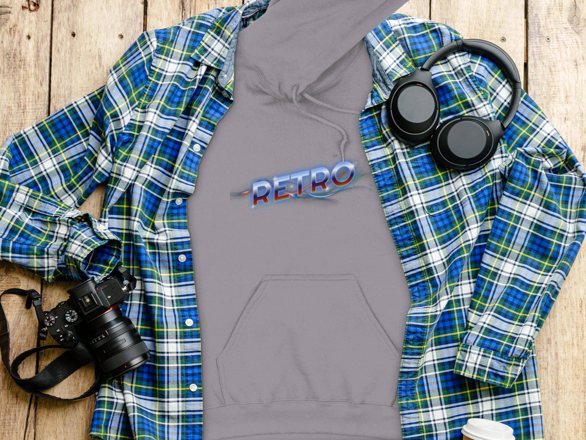 A flat lay features a Garment Graphics blue-green plaid shirt, a gray RETRO hoodie, black headphones, a camera, and a white travel mug on a wooden surface for a modern trendy look.