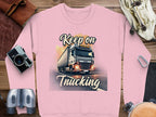 The Garment Graphics pink unisex sweatshirt showcases a truck at sunset with Keep on Trucking above and below, surrounded by travel essentials like a camera, binoculars, and leather bag.