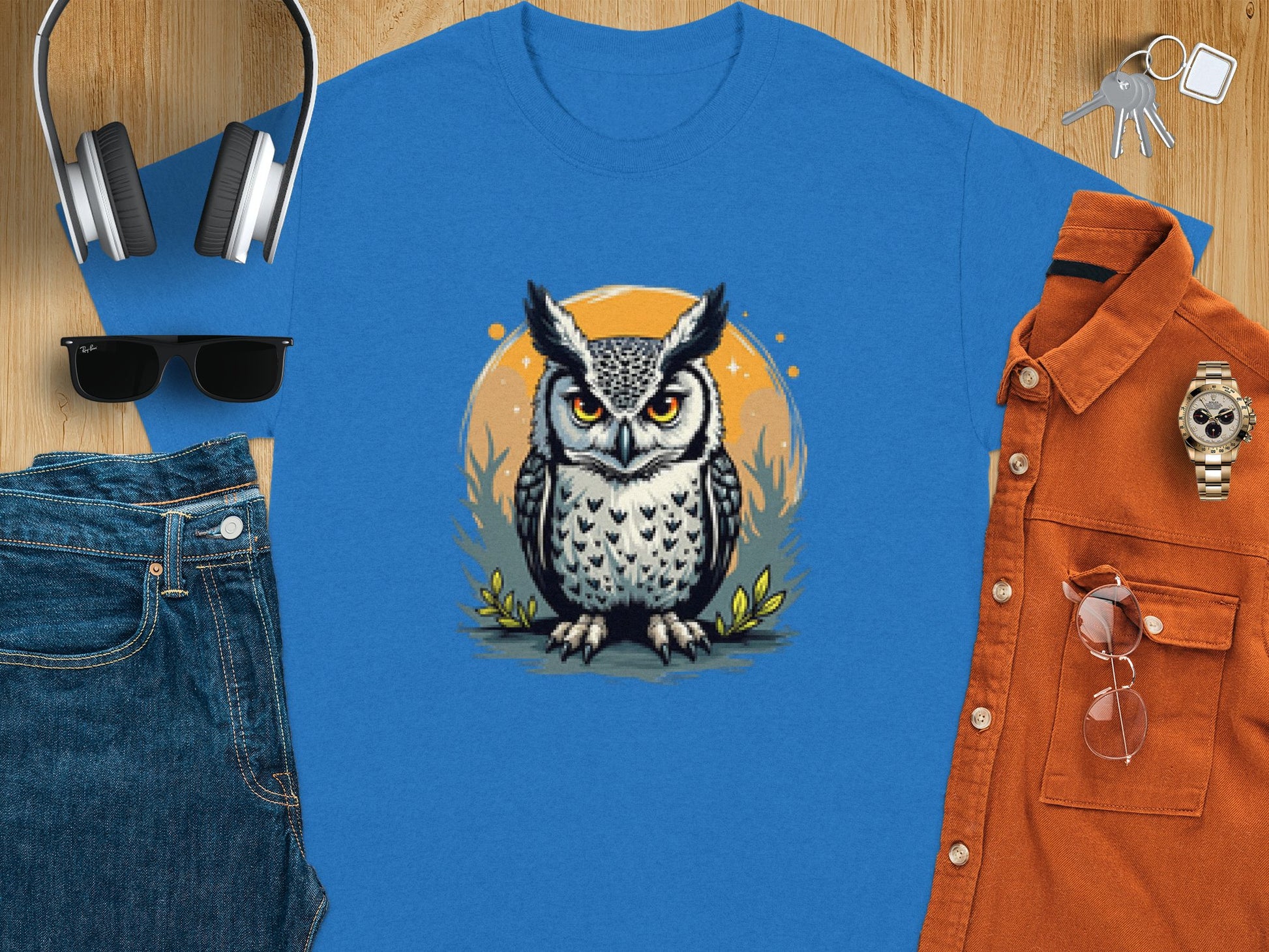 A flat lay features a Garment Graphics blue T-shirt with an owl graphic, paired with denim jeans, an orange button-up shirt, a watch, sunglasses, wireless headphones, and a set of keys on a wooden surface.