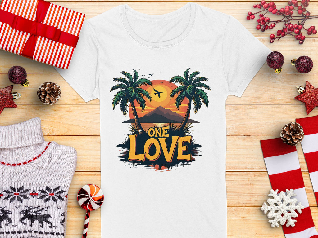 A Garment Graphics white T-shirt boasts a Tropical Sunset design with palm trees and One Love, ideal for beach outings. It sits among Christmas ornaments, a knitted sweater, candy, and wrapped gifts on a wooden surface.