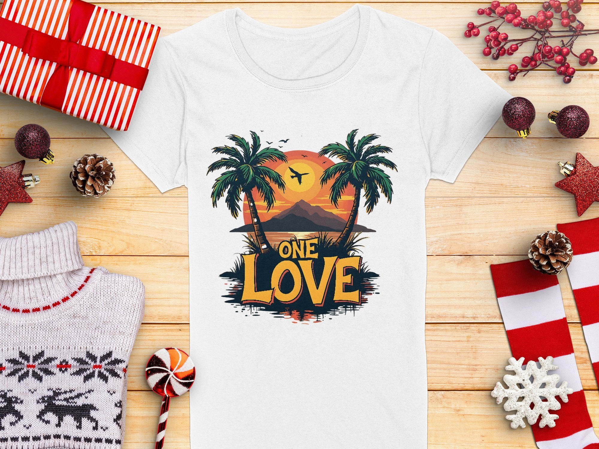 A Garment Graphics white T-shirt boasts a Tropical Sunset design with palm trees and One Love, ideal for beach outings. It sits among Christmas ornaments, a knitted sweater, candy, and wrapped gifts on a wooden surface.