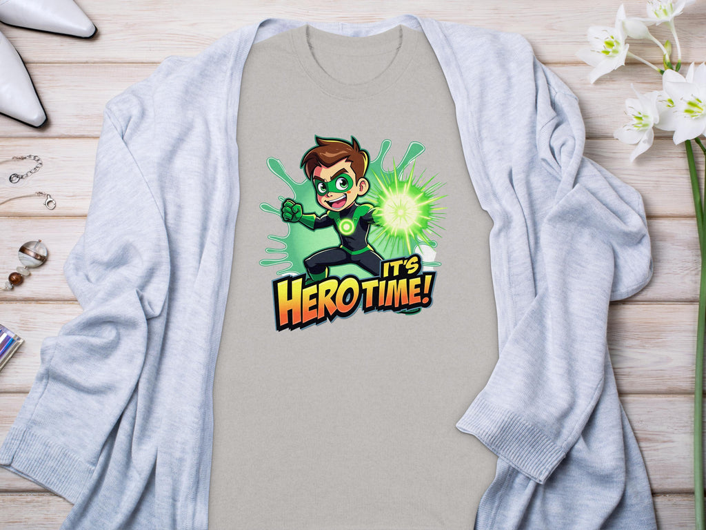 A vibrant light grey t-shirt from Garment Graphics with a Its Hero Time! superhero print lies on wood, paired with a matching cardigan, white heels, chic accessories, and delicate white flowers, setting the stage for stylish family sizing adventures.