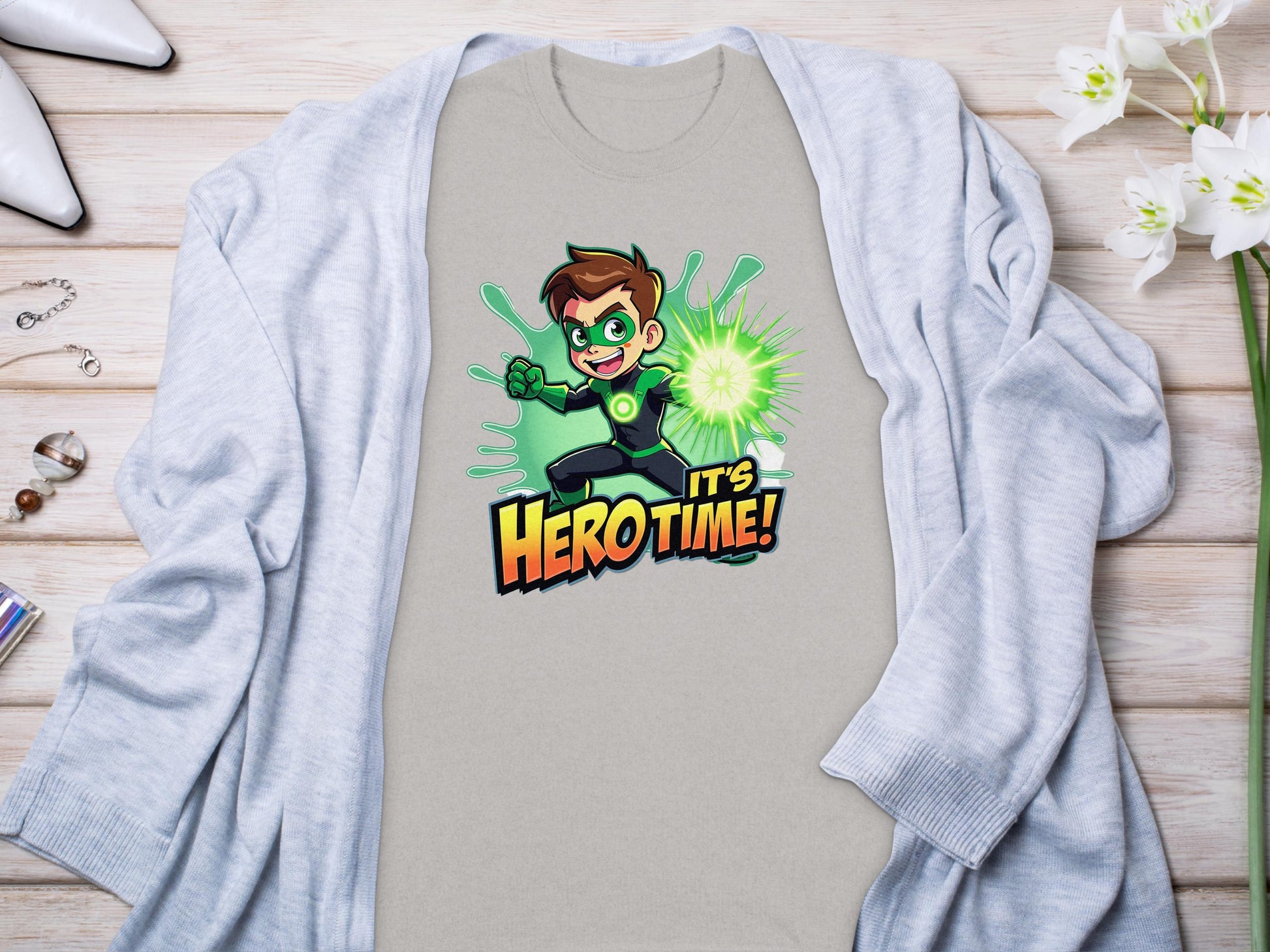 A vibrant light grey t-shirt from Garment Graphics with a Its Hero Time! superhero print lies on wood, paired with a matching cardigan, white heels, chic accessories, and delicate white flowers, setting the stage for stylish family sizing adventures.