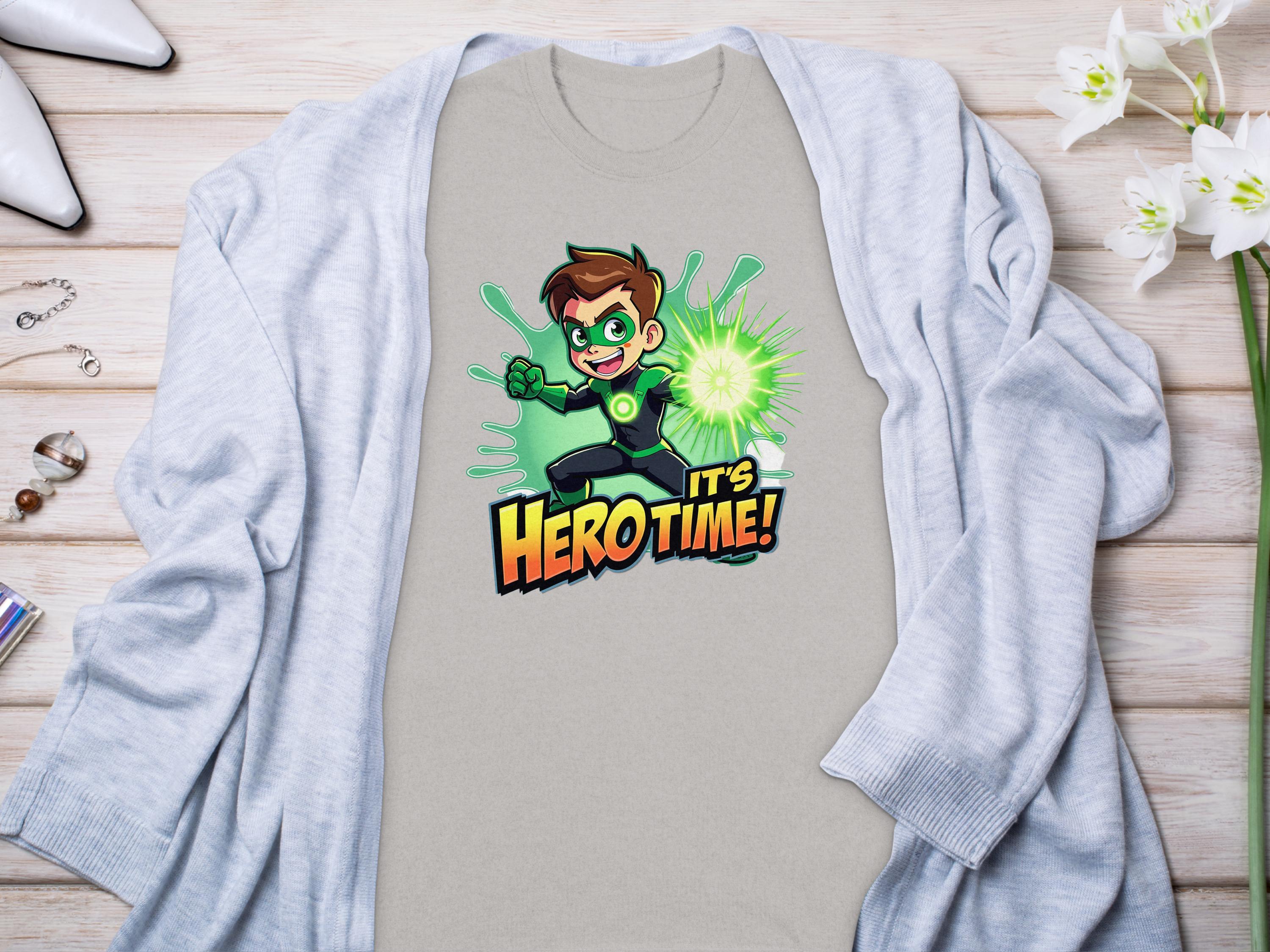 A vibrant light grey t-shirt from Garment Graphics with a Its Hero Time! superhero print lies on wood, paired with a matching cardigan, white heels, chic accessories, and delicate white flowers, setting the stage for stylish family sizing adventures.
