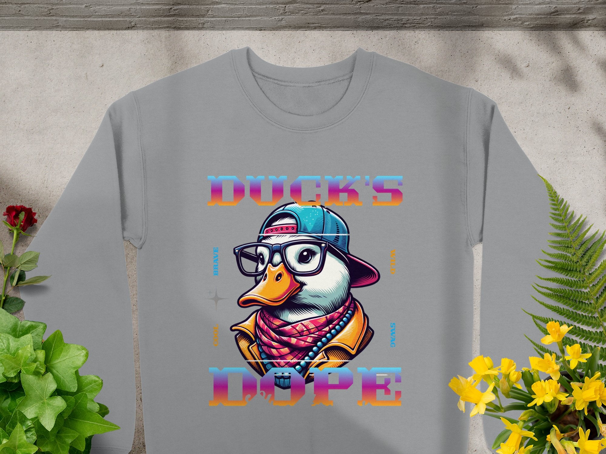 This trendy Garment Graphics sweatshirt has a gray base with a vibrant duck cartoon in glasses, a beanie, and a scarf. Featuring the bold text DUCKS DOPE, its styled against concrete amidst green and yellow plants.