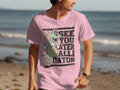 A man on the beach wears Garment Graphics casual wear—a pink T-shirt with a green alligator holding a ball, waving, and saying See You Later Alligator. The ocean is in the background.