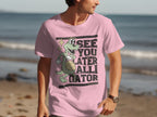 A man on the beach wears Garment Graphics casual wear—a pink T-shirt with a green alligator holding a ball, waving, and saying See You Later Alligator. The ocean is in the background.