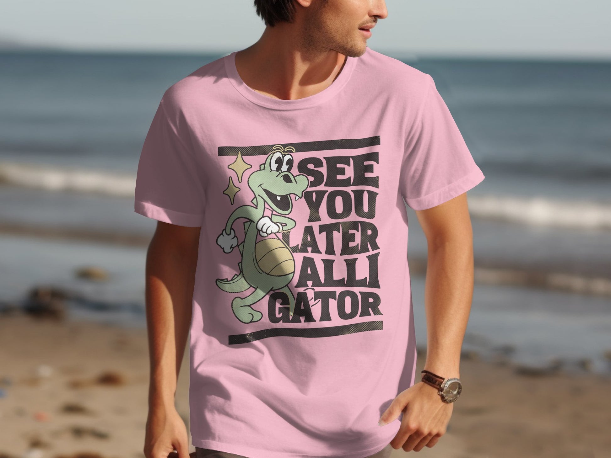 A man on the beach wears Garment Graphics casual wear—a pink T-shirt with a green alligator holding a ball, waving, and saying See You Later Alligator. The ocean is in the background.